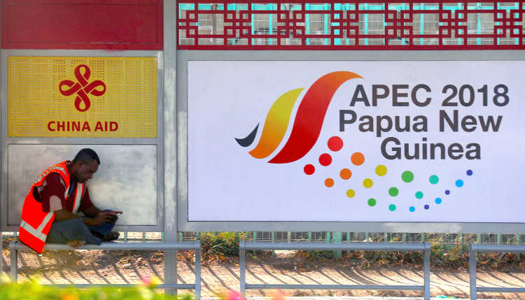 A man in a high-visibility waistcoat sits in the shadow of posters of China Aid and APEC 2018 Papua New Guinea