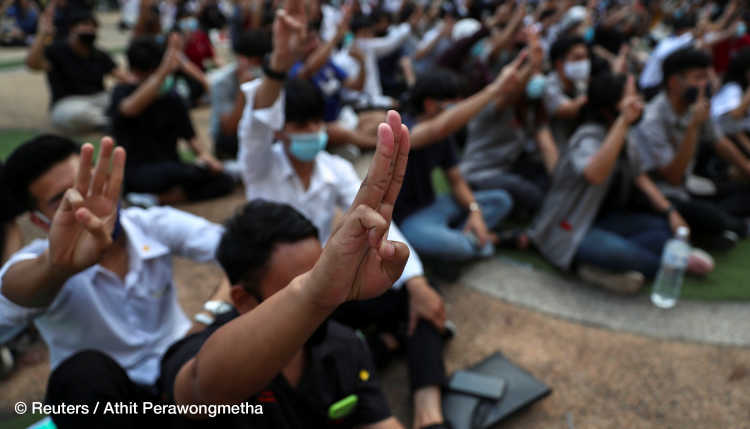 Protesters in Bangkok sit on the ground and hold up three fingers.