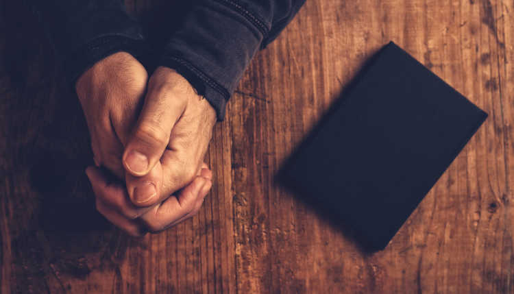 Verschränkte Hände auf einem Tisch neben einem Gebetsbuch.