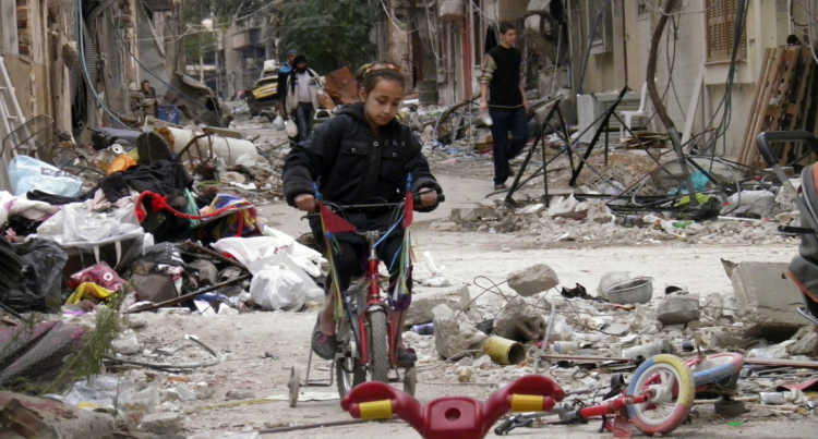 A Syrian girl rides a child's bicycle with training wheels through a street littered with rubble.