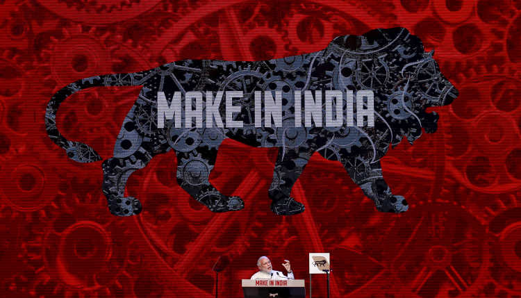 Prime Minister Modi sits in front of a large poster promoting investment ("Make in India").